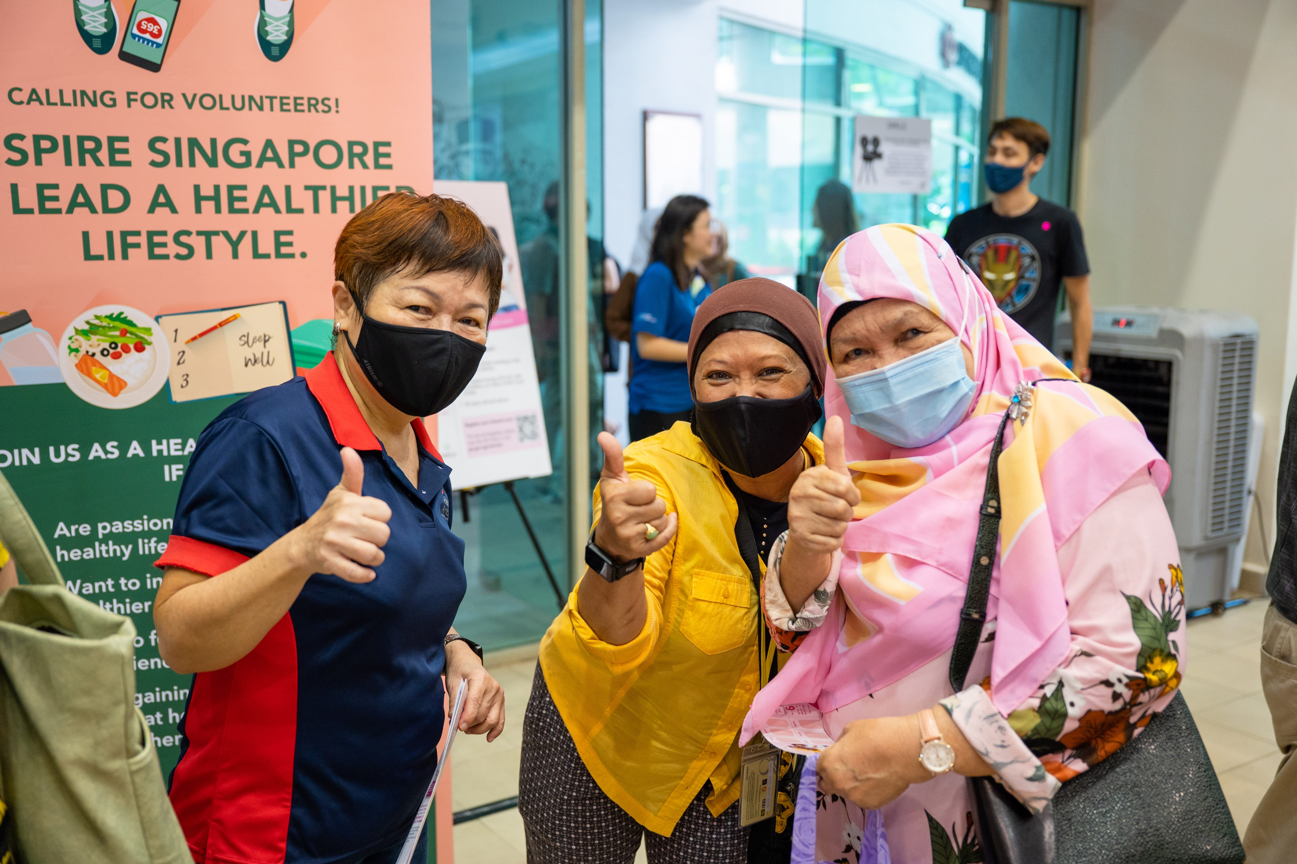 Three women giving thumbs-up at a community health outreach event, standing beside a volunteer recruitment banner promoting healthier lifestyles.